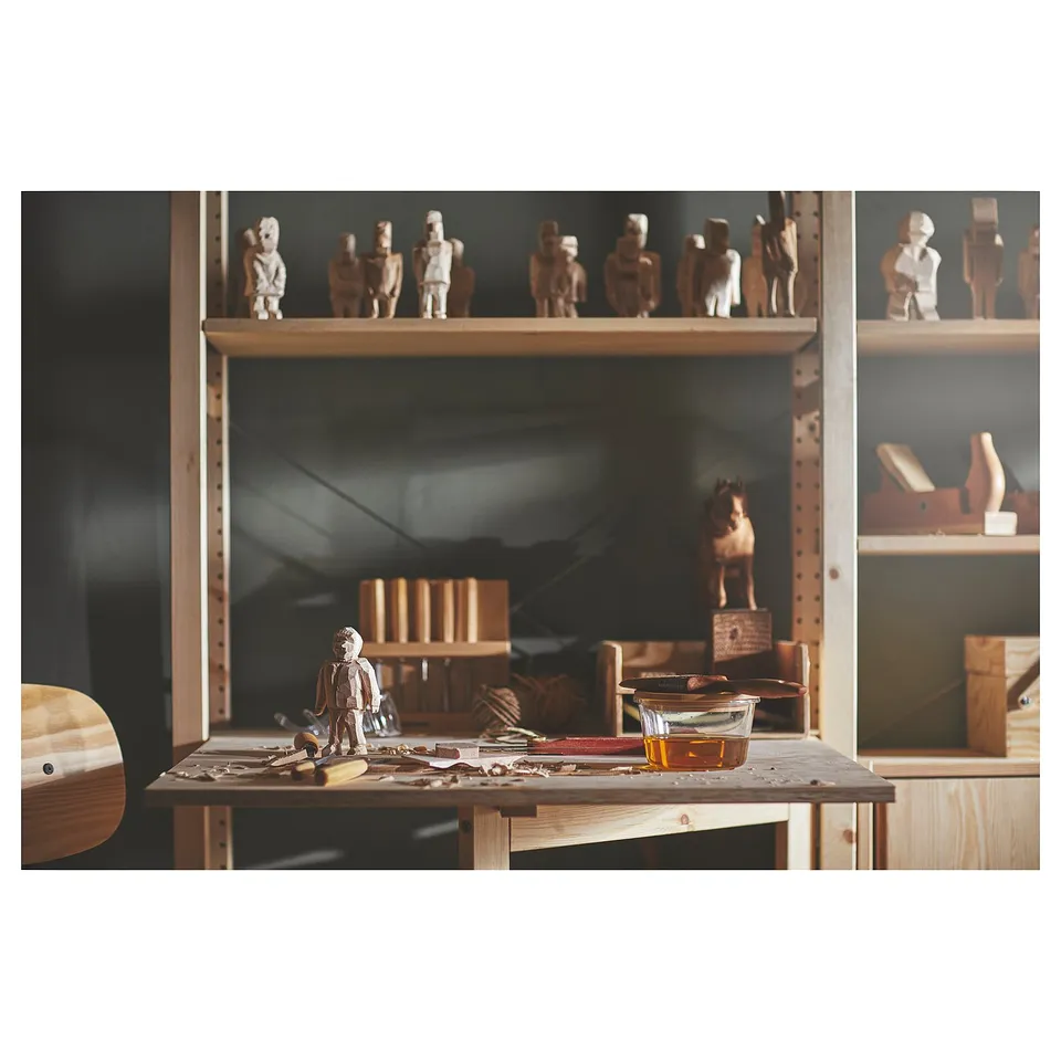 Workshop with wooden carvings. Shelves hold human figurines, tools, and a bowl on a wooden table. Dark background.