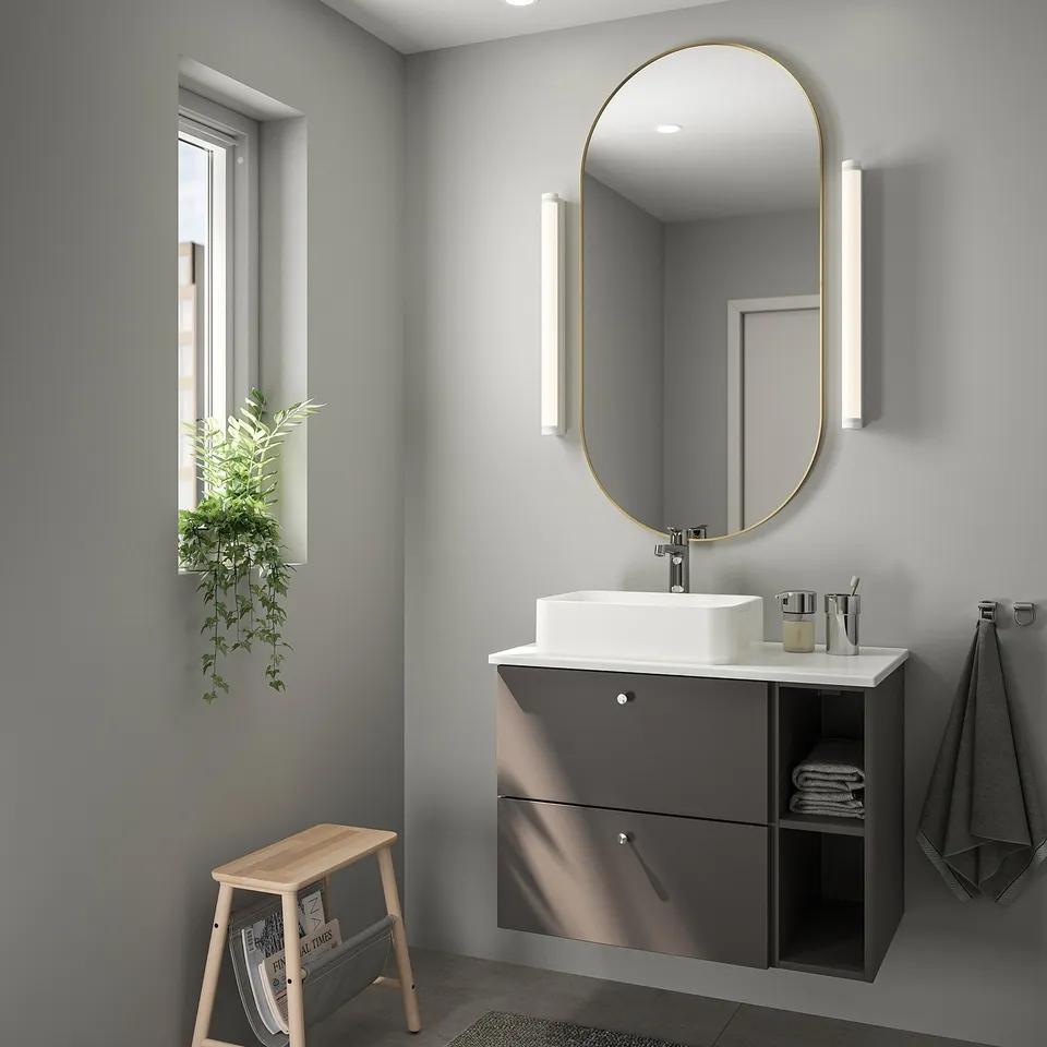 Modern bathroom with large gold-framed mirror, dark grey floating vanity, wooden stool, plant, and grey towels.