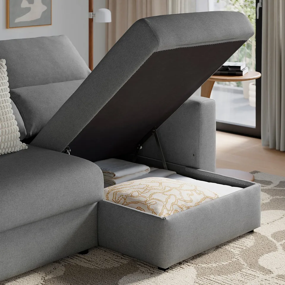 Grey sofa with lifted storage seat revealing cushions and blankets inside.