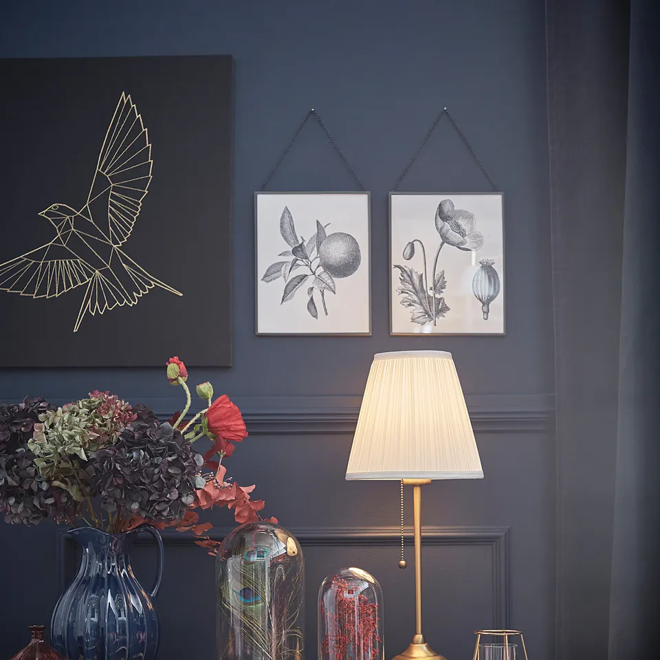 Dark blue wall with art, lamp, flowers, and decor. Golden ÅRSTID lamp has fabric shade, brass base.