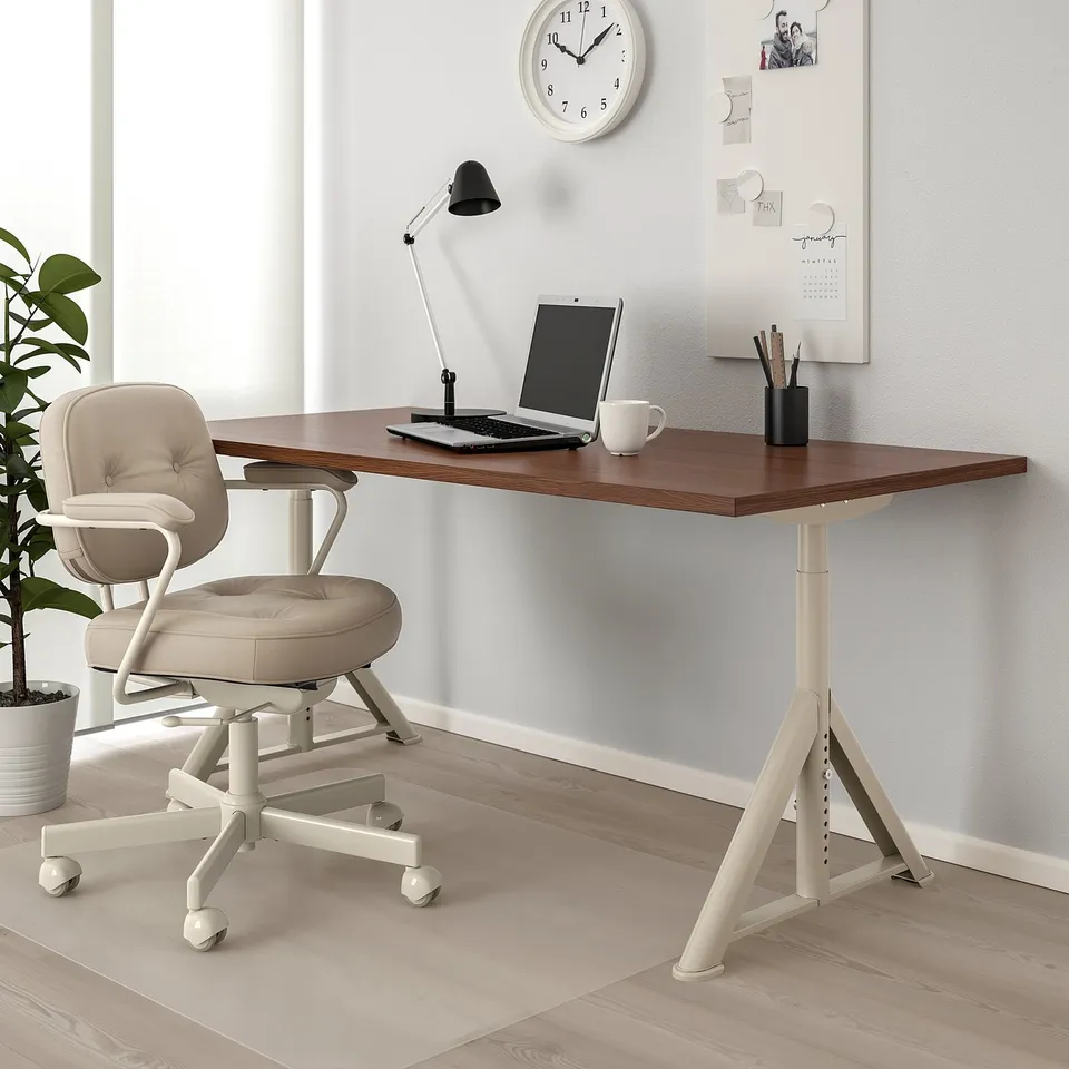IDÅSEN desk in minimalist office with wooden top, off-white legs, cable management, and a beige chair underneath. Clock and plant nearby.