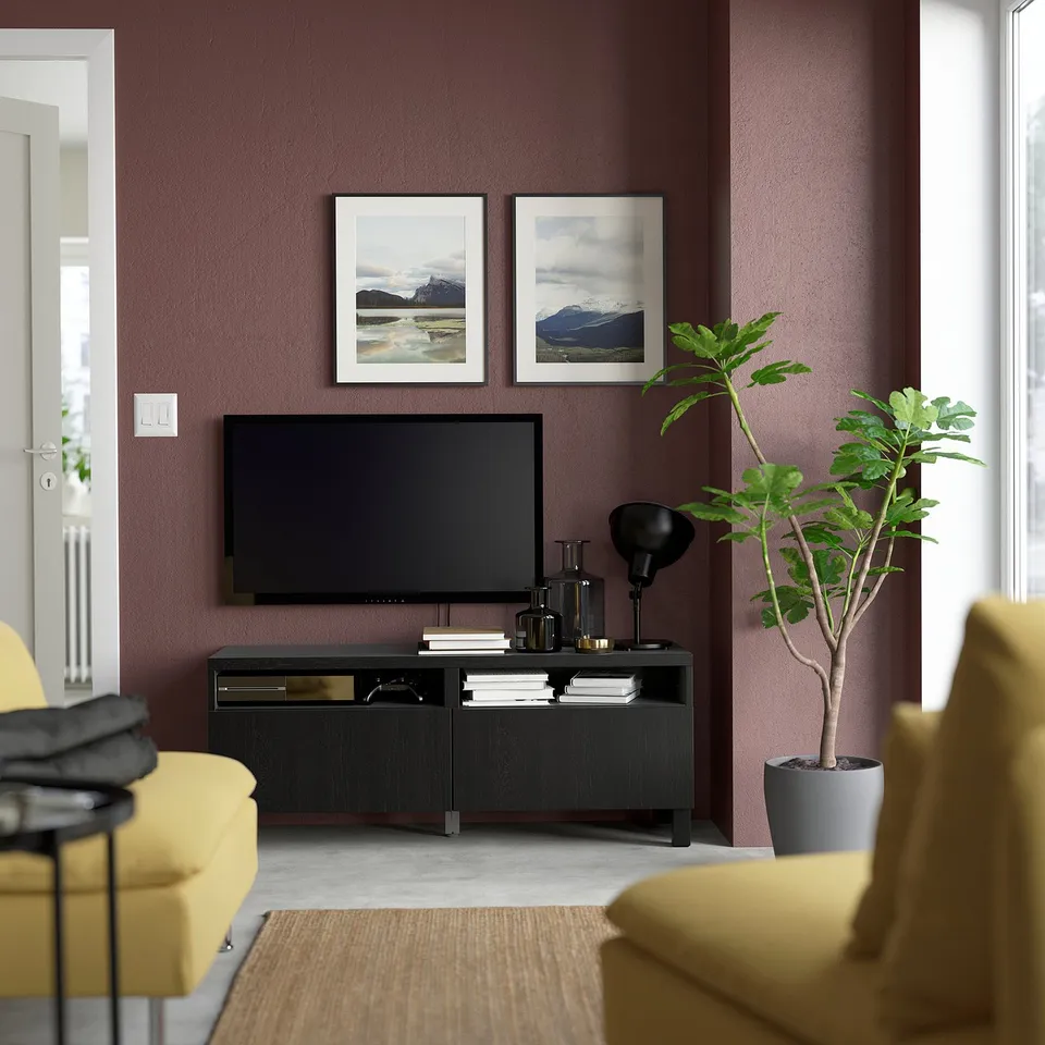 Living room with tv and yellow couch. BESTÅ tv stand in black, mounted on purple wall. Two framed nature pictures above, tall plant to right.