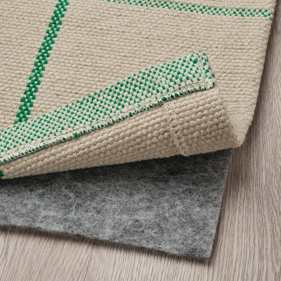 Off-white rug with green border and dots, folded back to show soft underside and grey felted non-slip pad.
