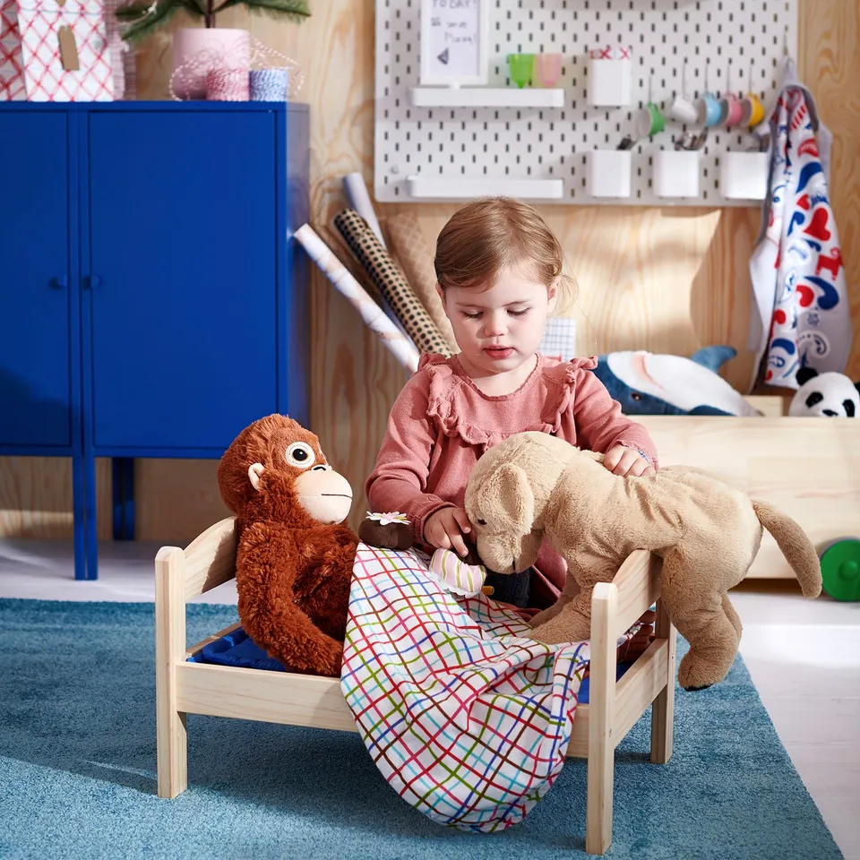 Little girl playing with soft brown monkey toy on small wooden chair. Colourful blanket and blue cabinet.