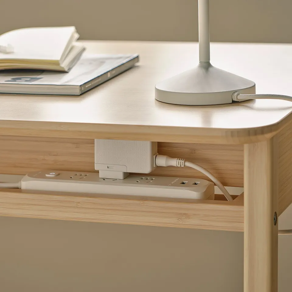 A wooden desk with cable management and a drawer. A lamp and books are on top.