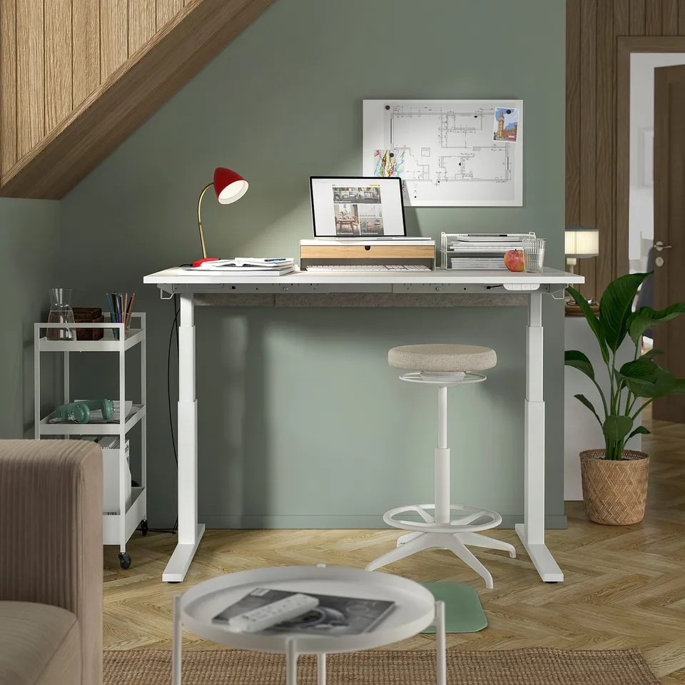 Modern home office with MITTZON white sit-stand desk, ergonomic stool, laptop, red lamp, plant, and organised storage.