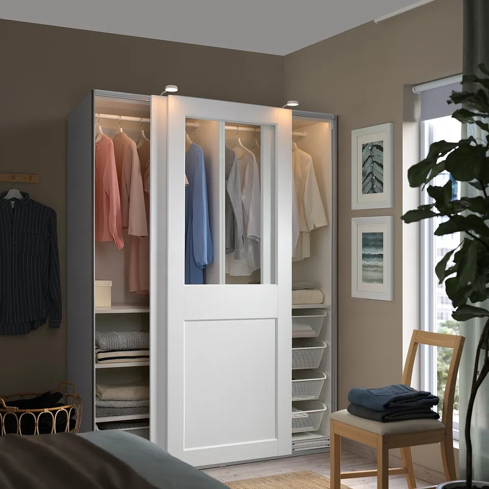 A white PAX wardrobe with glass doors displaying clothes neatly. It stands tall beside a window, larger than a nearby chair, with art on the wall.