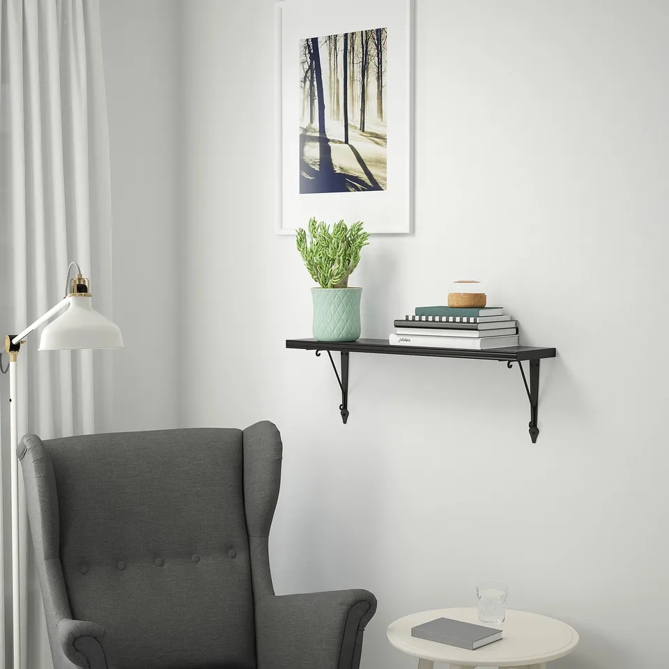 Calm room with grey armchair, small white side table, black shelf holding books, plant, and decor under framed forest picture.