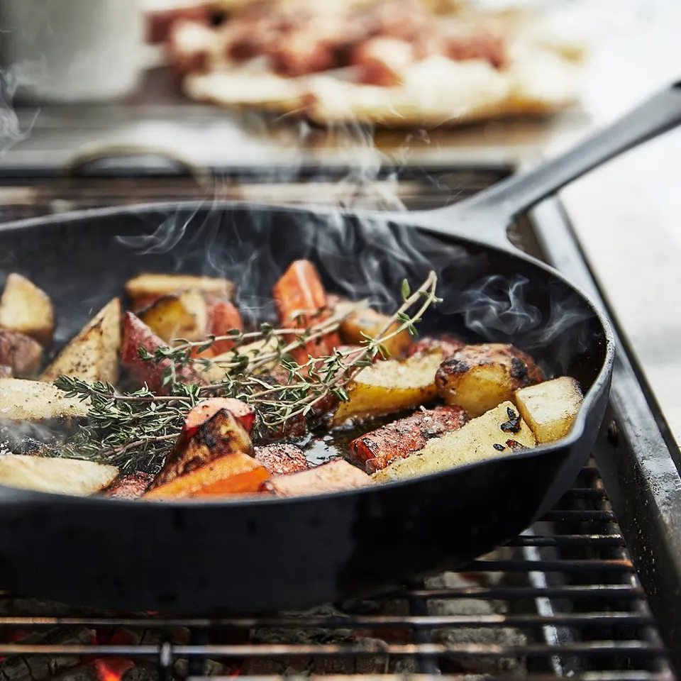 Black cast-iron skillet grilling colourful vegetables and herbs on a modern black grill with smoke rising.