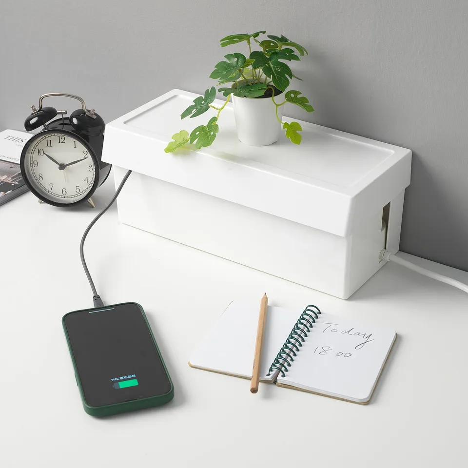 Clean desk with white SÄTTING box. Hides cables, charges phone. Alarm clock, plant, notes nearby.