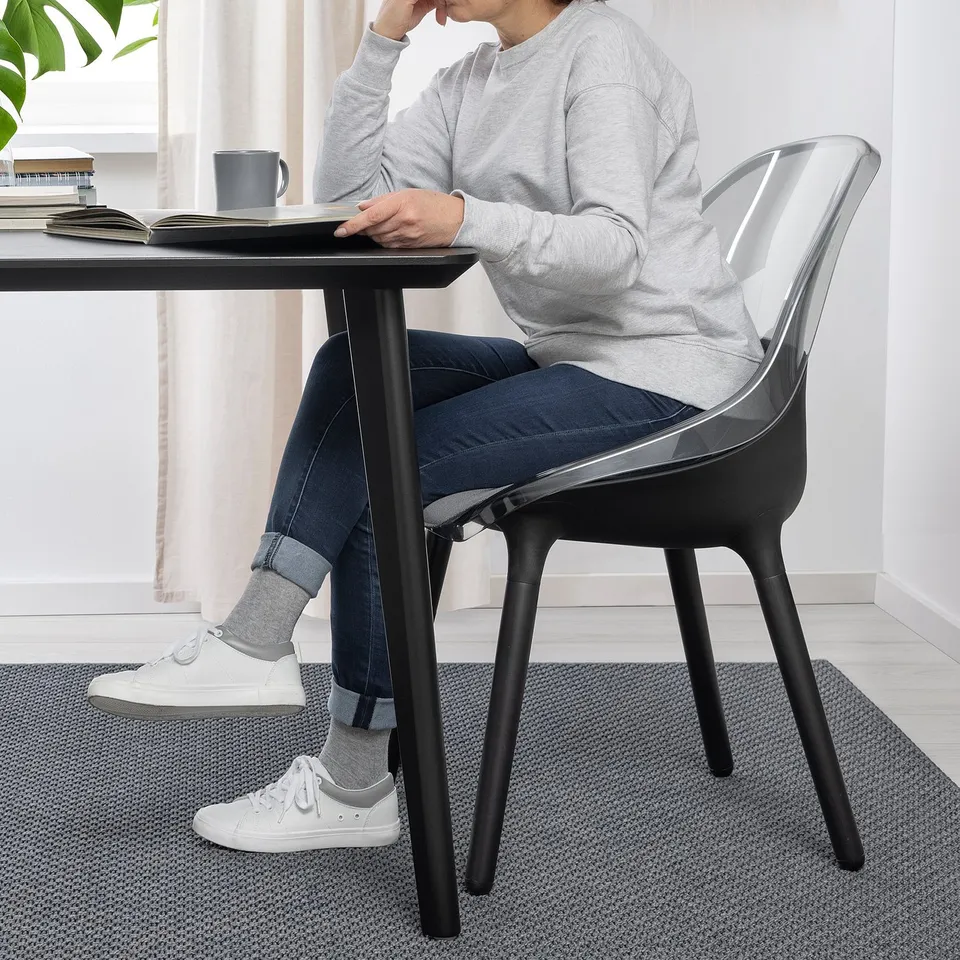 Person sitting at modern dining table on BALTSAR chair with transparent seat, sturdy beech legs. Chair offers comfort and stability.