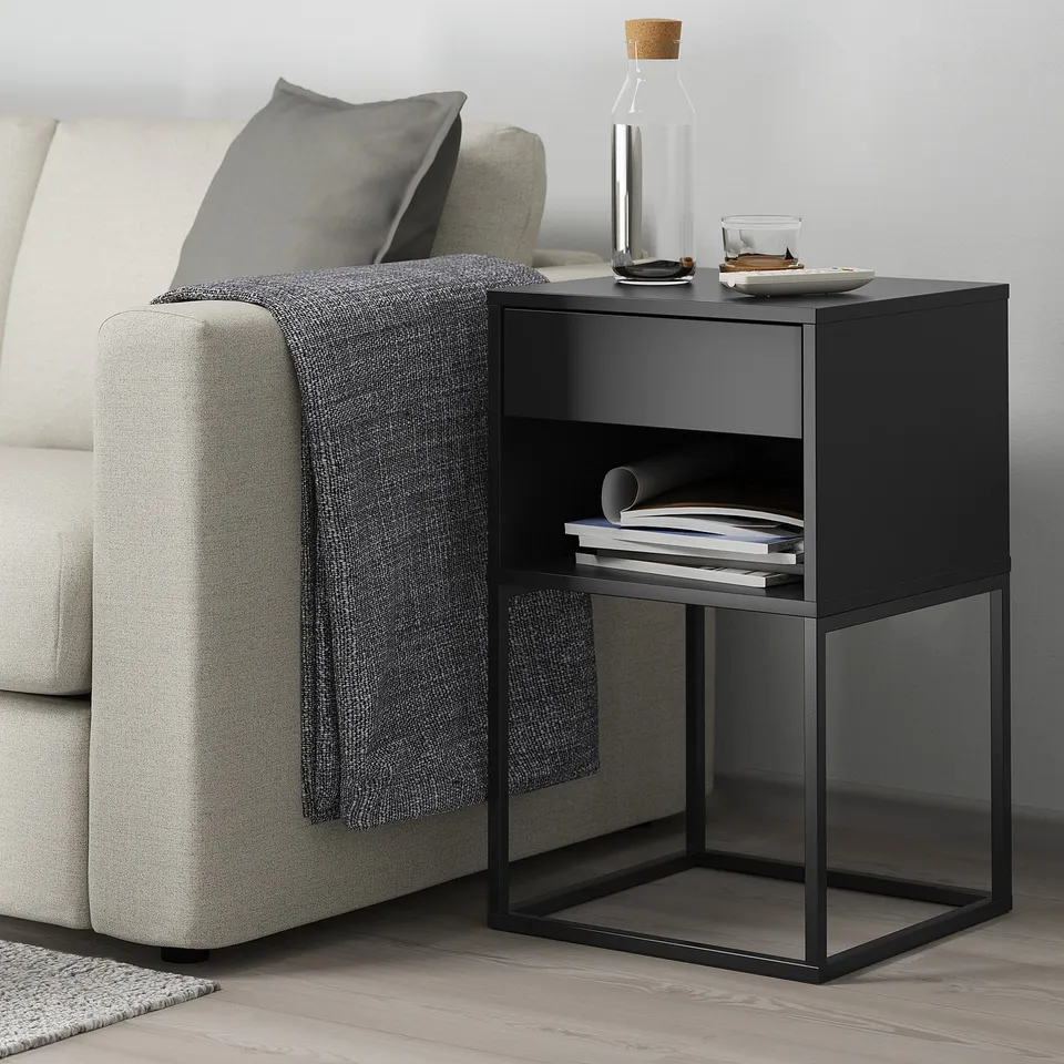 Vs: a black VIKHAMMER side table next to a grey sofa. It is compact, with two drawers and an open shelf, holding books and decor items.