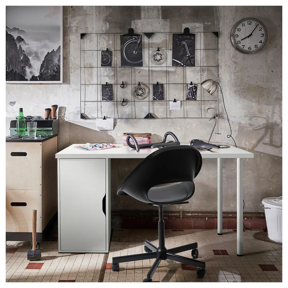 Industrial-style desk setup: black chair, white desk, grey wall with clock and art, black lamp.