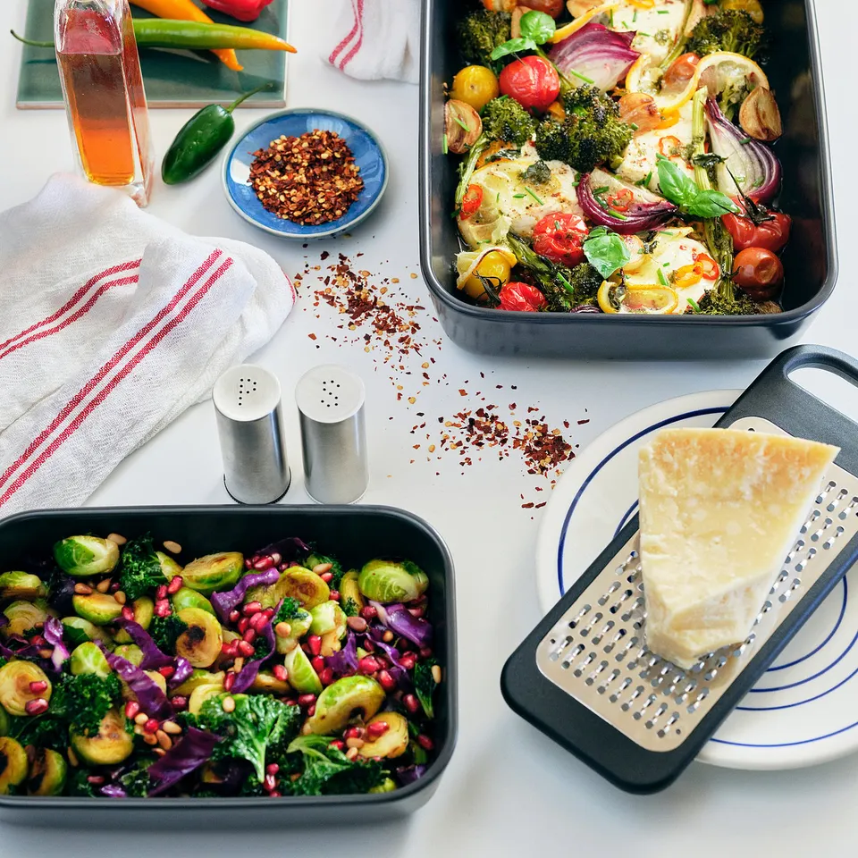 A table with various dishes: a baking tray with vegetables, a grater with cheese, and a salad bowl.