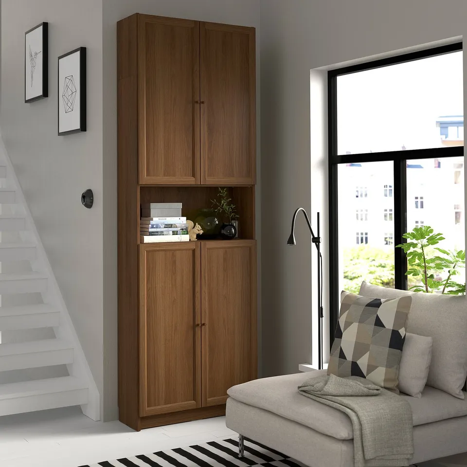 Wooden BILLY bookcase in corner, tall and slim with open and closed storage, near large window and grey sofa.
