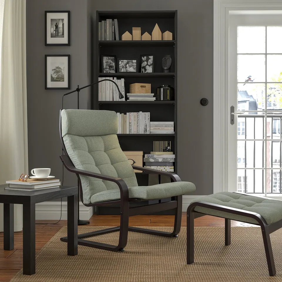 Modern reading nook with POÄNG chair & footstool in light green fabric, dark wood frame, cushioned seat & back. Near a large bookshelf & window.