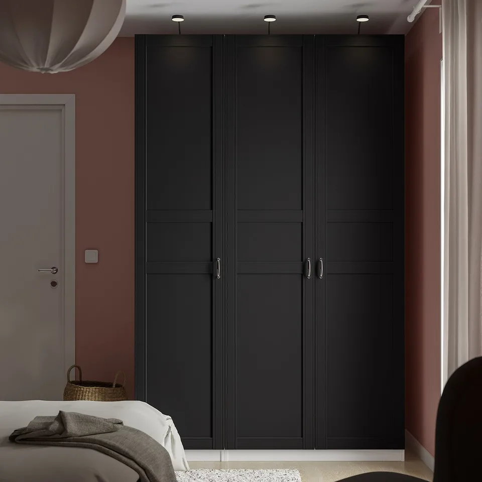 Modern bedroom with large black PAX wardrobe, next to a white door, partially covering a bed with grey blankets. Wardrobe is tall and narrow.