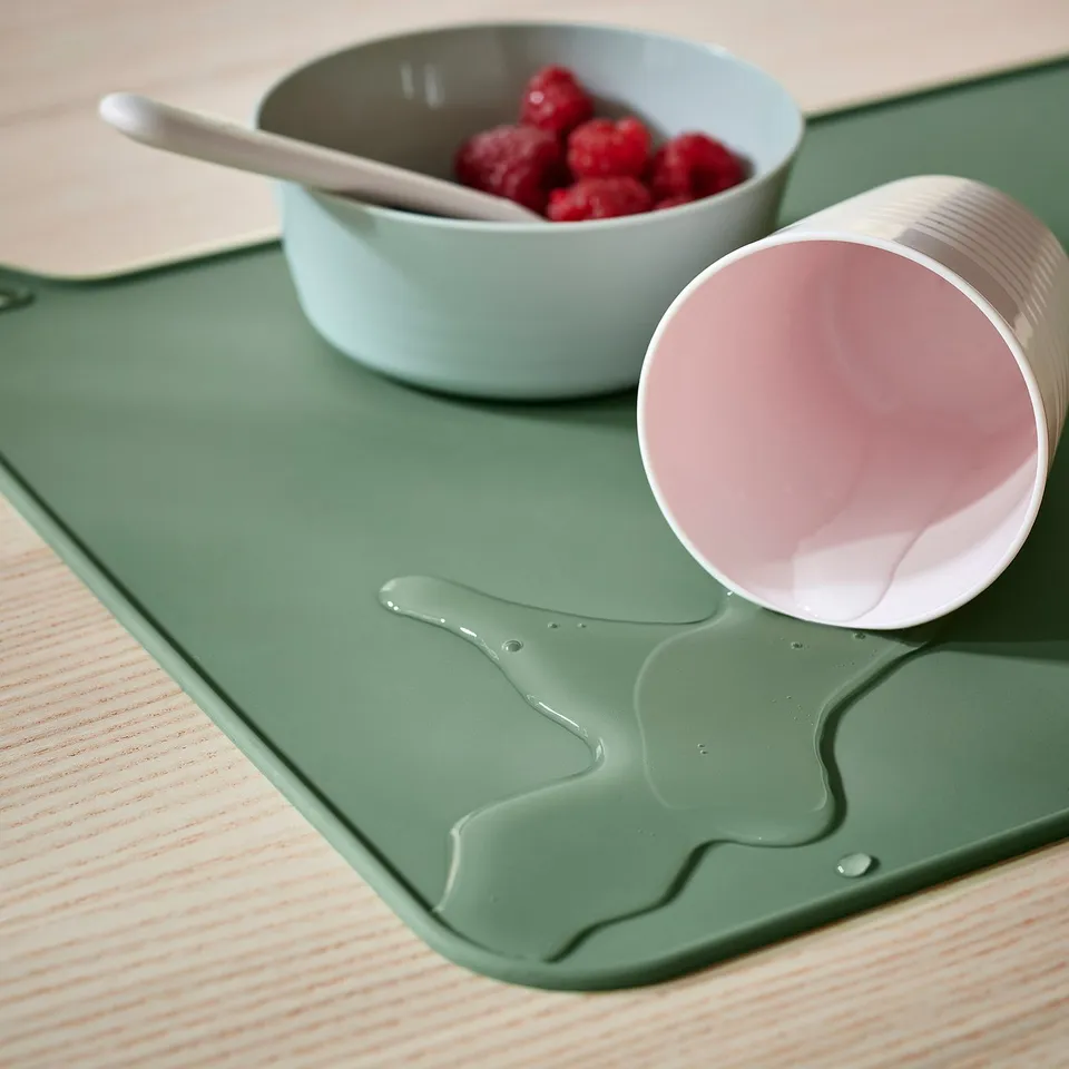 Yoghurt container spills on green cat-shaped silicone place mat with bowl of raspberries.