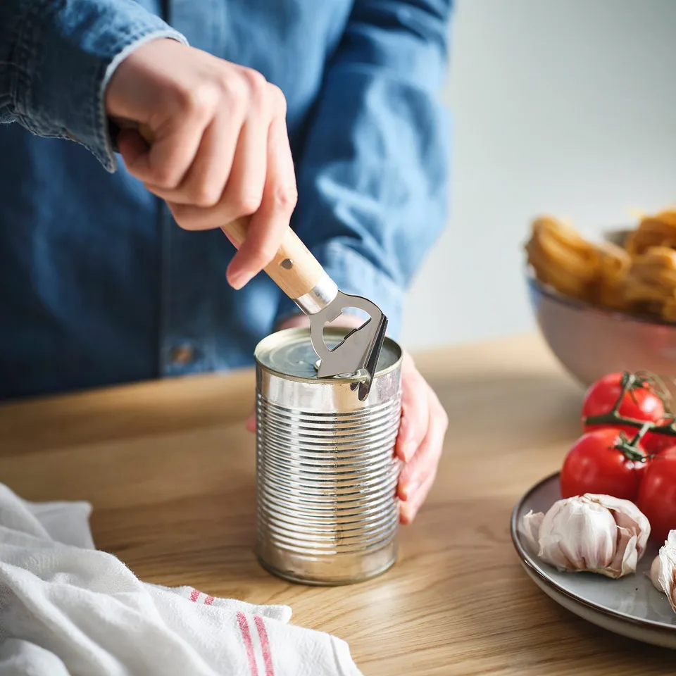 Person opens tin can using VARDAGEN bottle/can opener with wooden handle. Fresh vegetables and kitchen utensils are on the table.