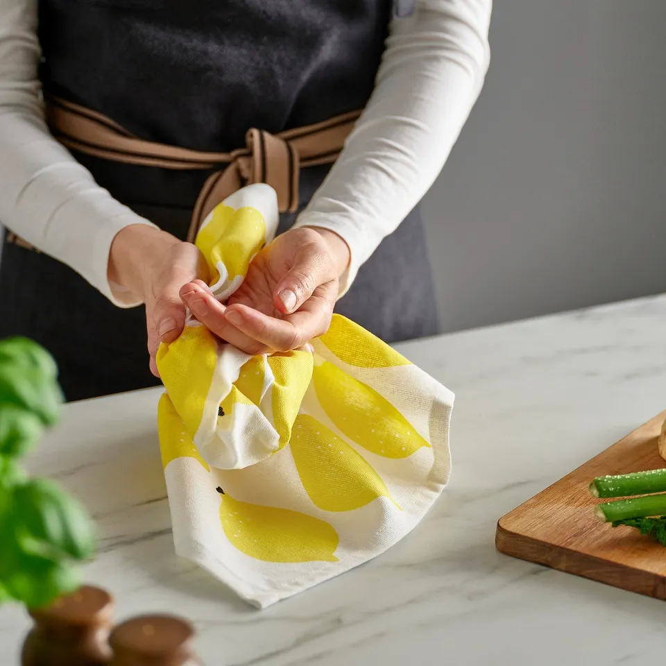 Drying hands with yellow towel. Counter has fresh herbs, spoons, and sliced cucumbers.