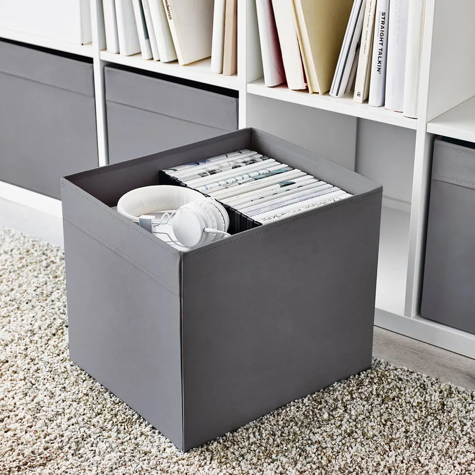 Grey storage bin on carpet next to bookshelf, holding folders and headphones.