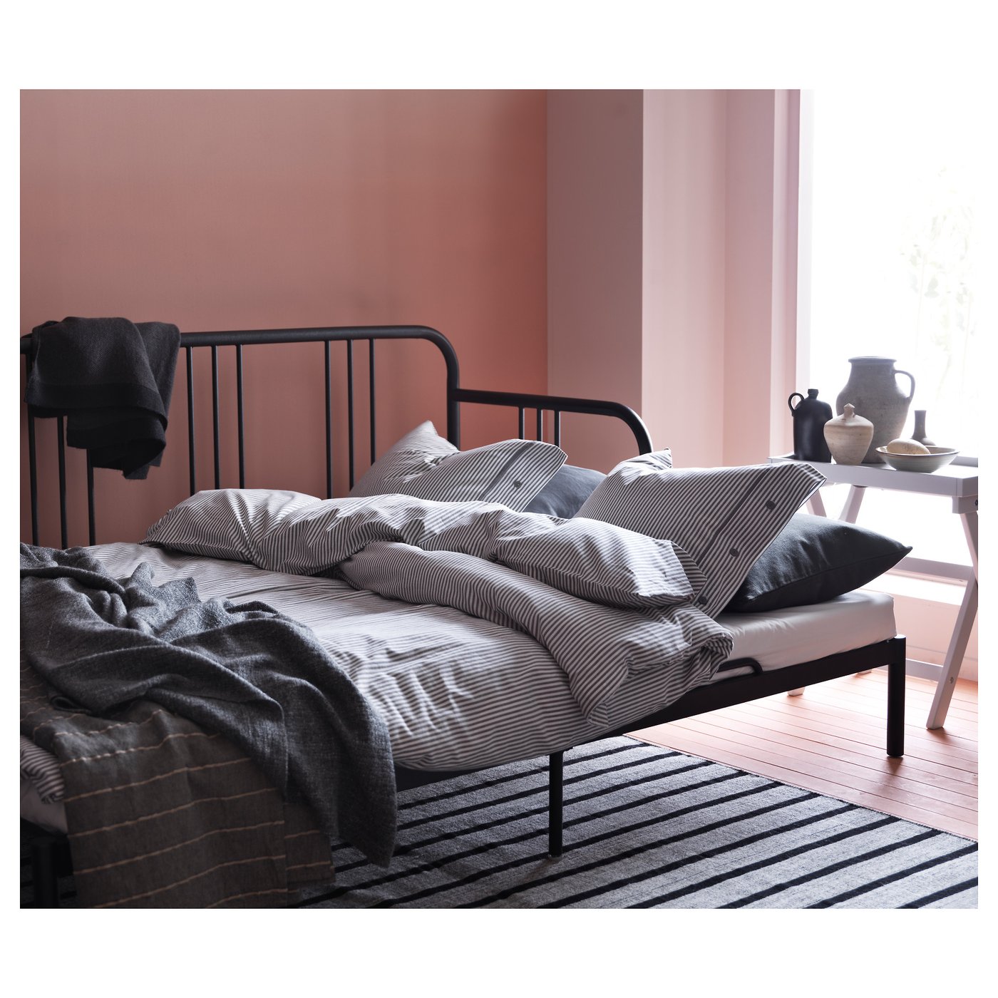 A FYRESDAL black metal day-bed with striped linens in a pink room, featuring a foam mattress, pillows, and decorations.