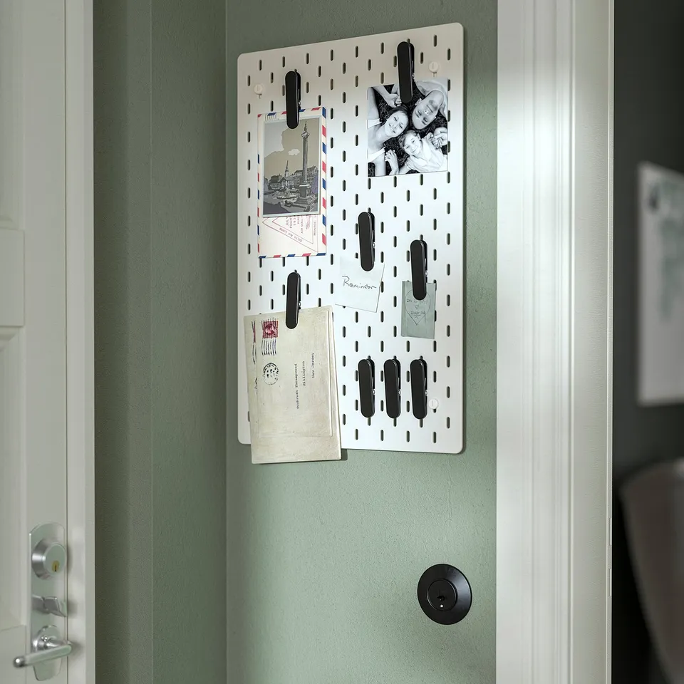 White SKÅDIS pegboard on green wall holds photos, postcards, and notes with black clips. Door with doorknob below.