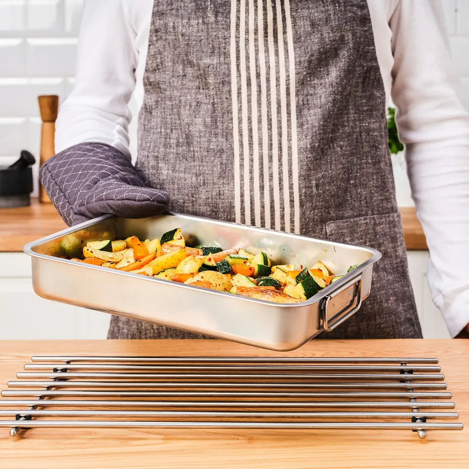 Person holding a metallic baking tray filled with colourful roasted vegetables on a wooden kitchen counter.