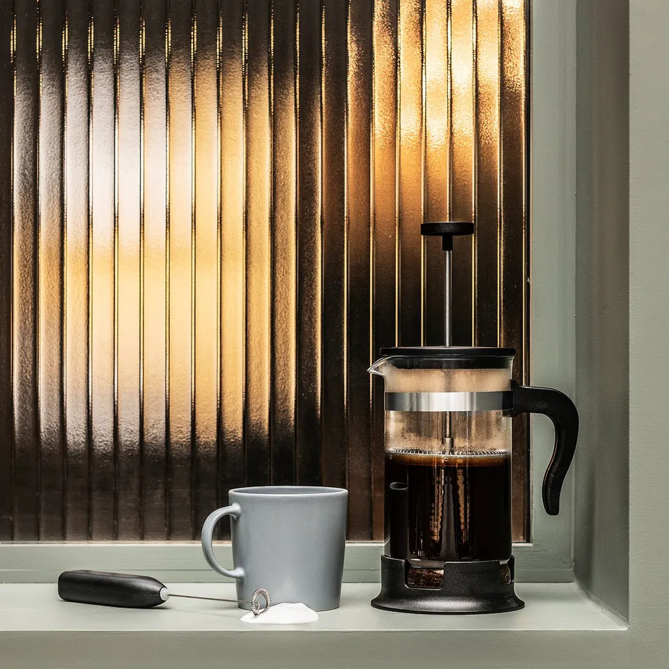 Black cafetière on window sill, grey cup, frother, sugar. Sunlight streams through blinds.
