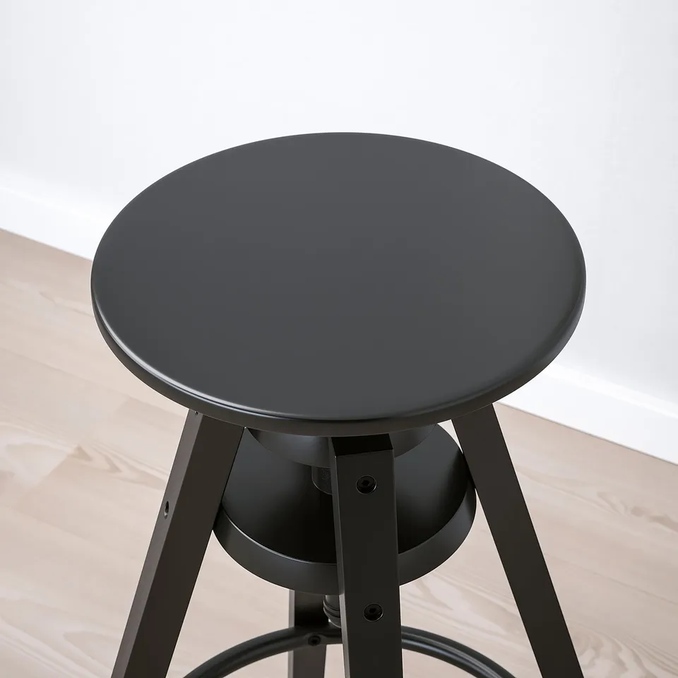 Black round stool with adjustable height, three steel legs, on wooden floor.