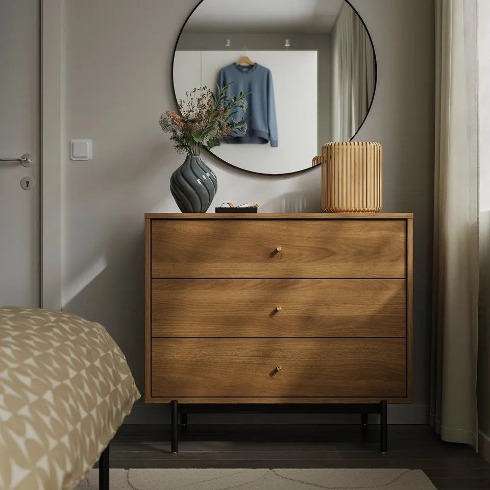 Wooden dresser RÅDMANSÖ with 3 brass knobs, dark top, next to bed, reflected in mirror, walnut tone contrasts bed sheets.