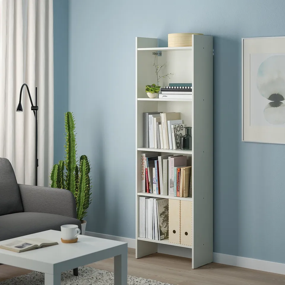 Modern white bookcase in living room, holding books & decor. Between grey chair and small table.