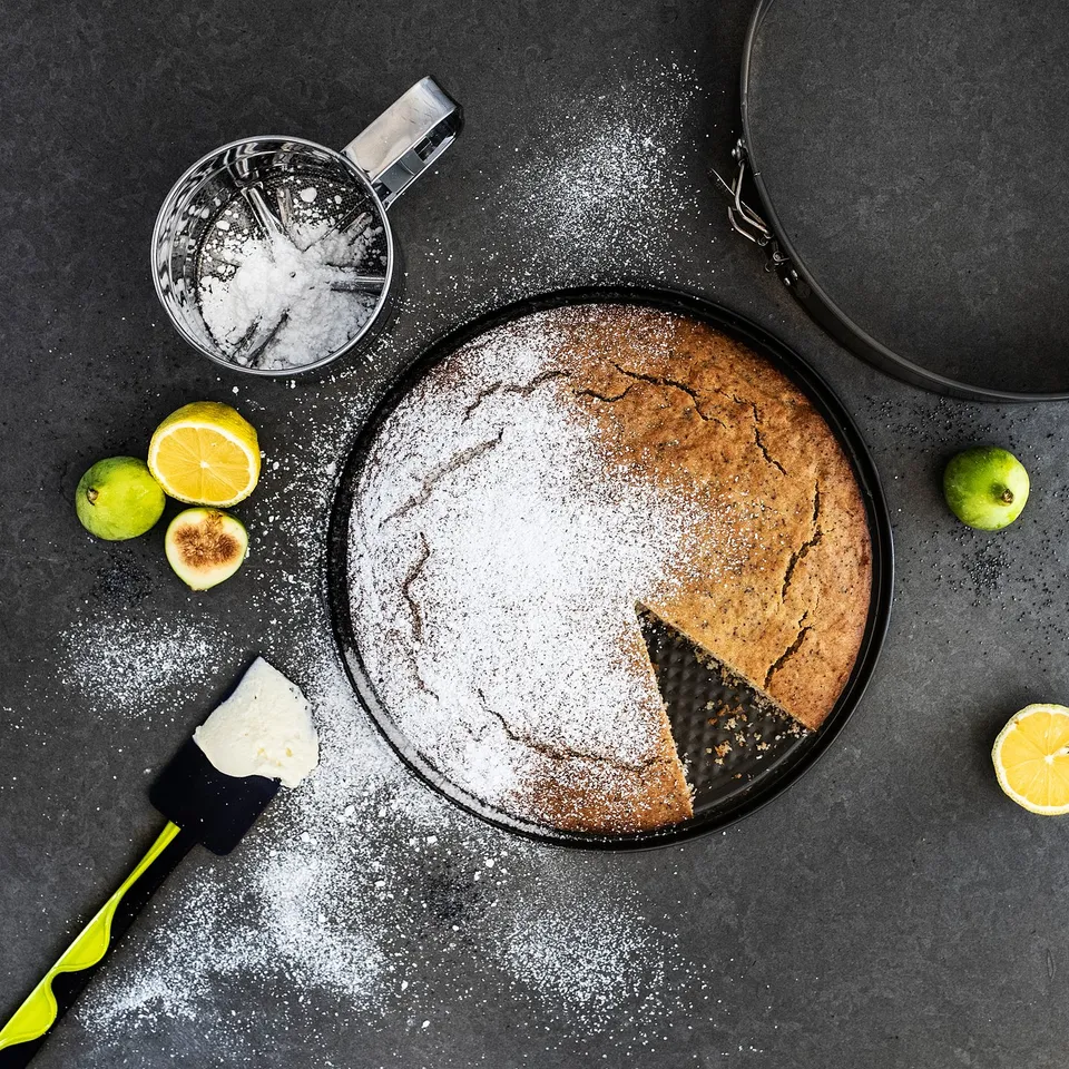 A golden cake on dark background, dusted with sugar, slice revealed, with lemons, apple, and baking tools.