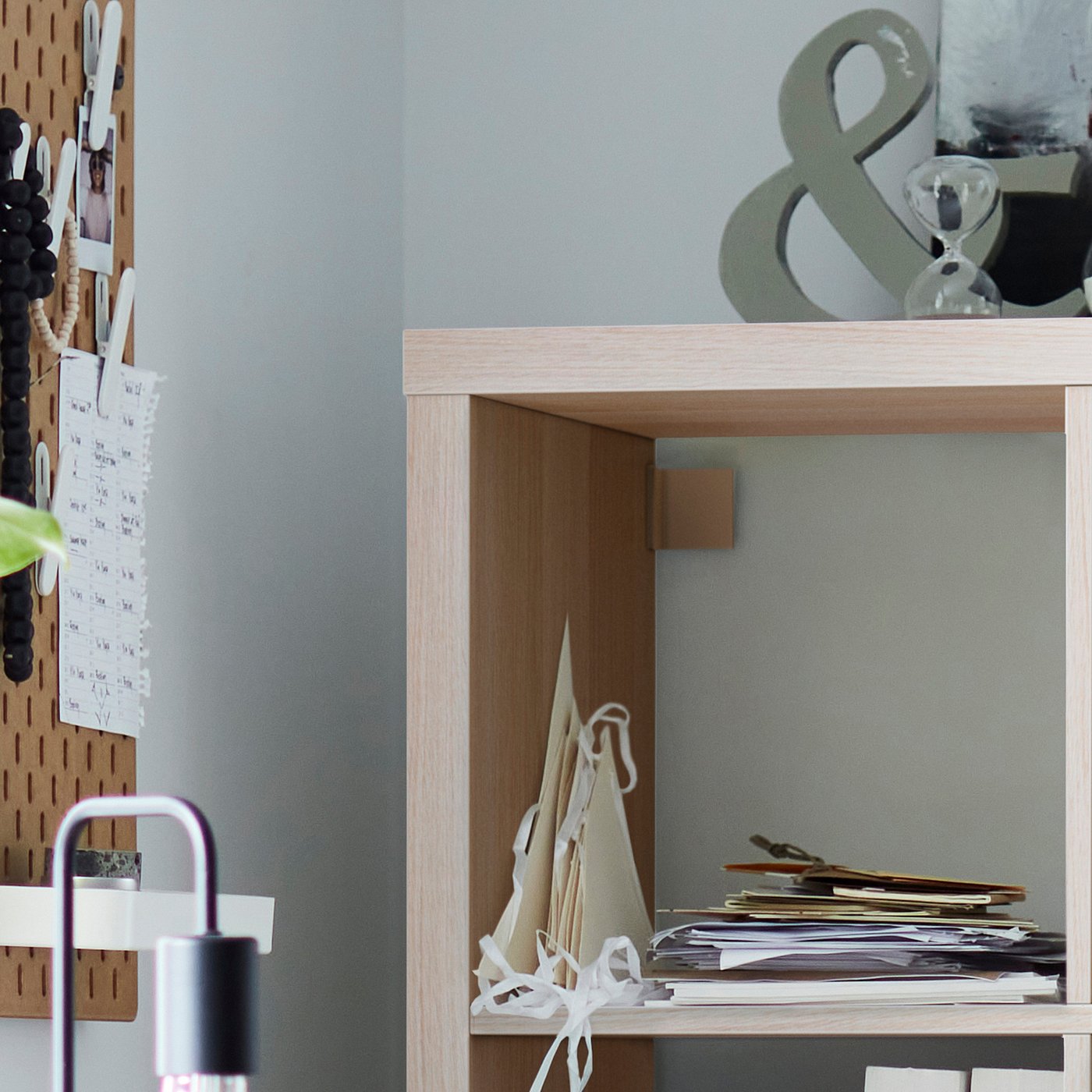Light wooden KALLAX shelf, empty top shelf, paper pile on bottom, grey wall art behind, pegboard with notes to left.
