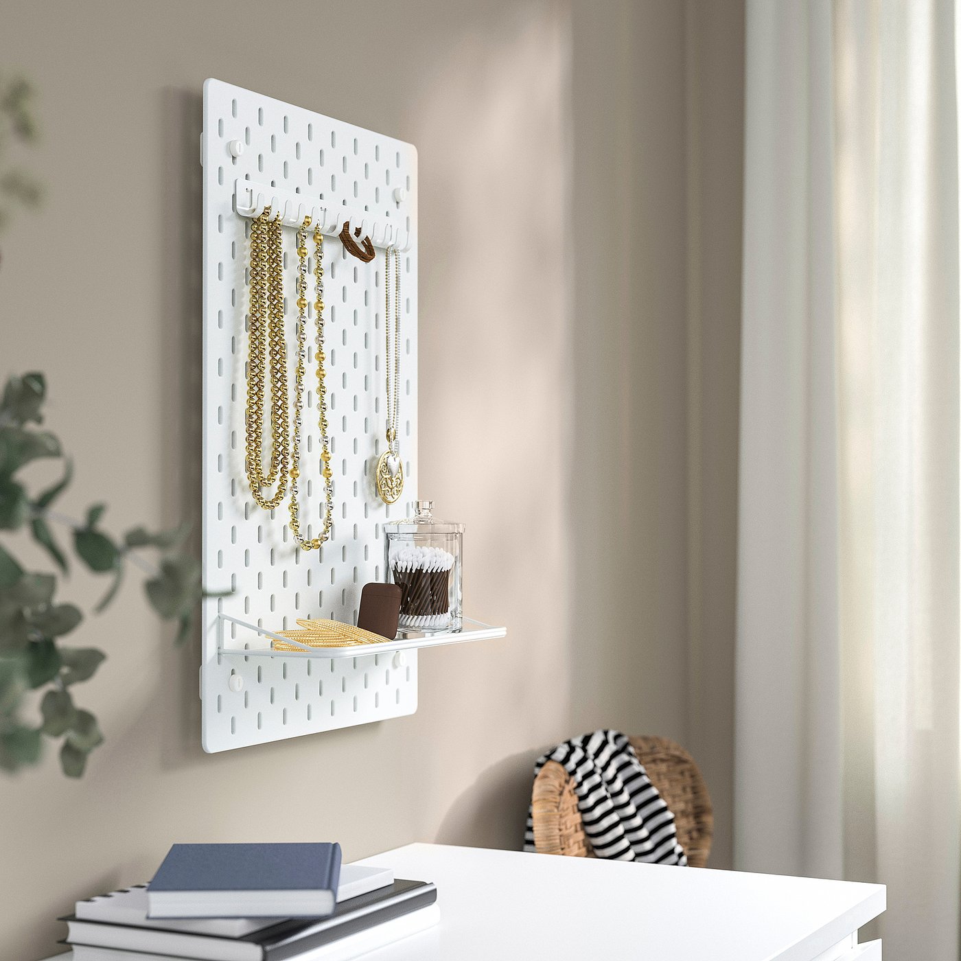 White SKÅDIS pegboard with hooks holding necklaces, shelf with container and wallet, near window with curtains.