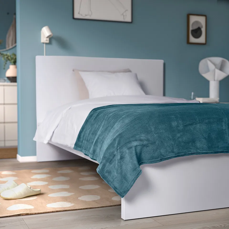 Cosy TRATTVIVA teal bedspread on a neat bed, in a calming blue & white room with a patterned rug.