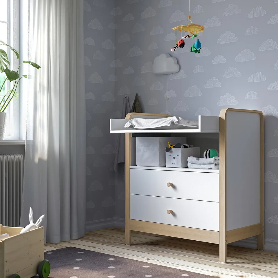 Nursery with ÄLSKVÄRD changing table, chest, and crib. Cloud-patterned wall, white curtain, mobile. Cosy and organised.