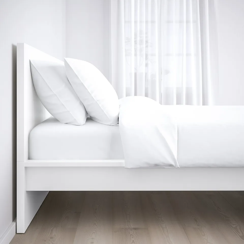 Clean, white bed with sleek headboard and two pillows, set against a bright window with sheer curtains, on a dark wooden floor.