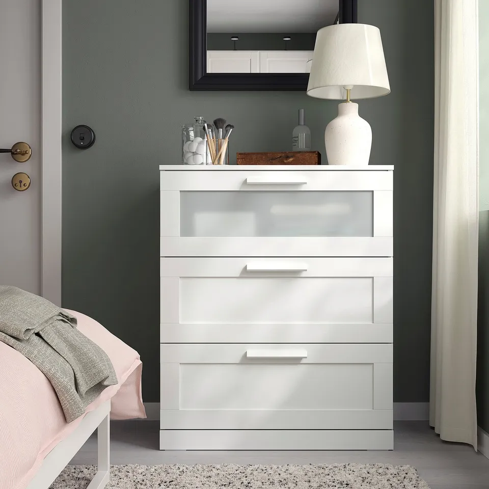 White BRIMNES chest with frosted glass top drawer, positioned between bed and window. It has four drawers, with a tall lamp and mirror above.