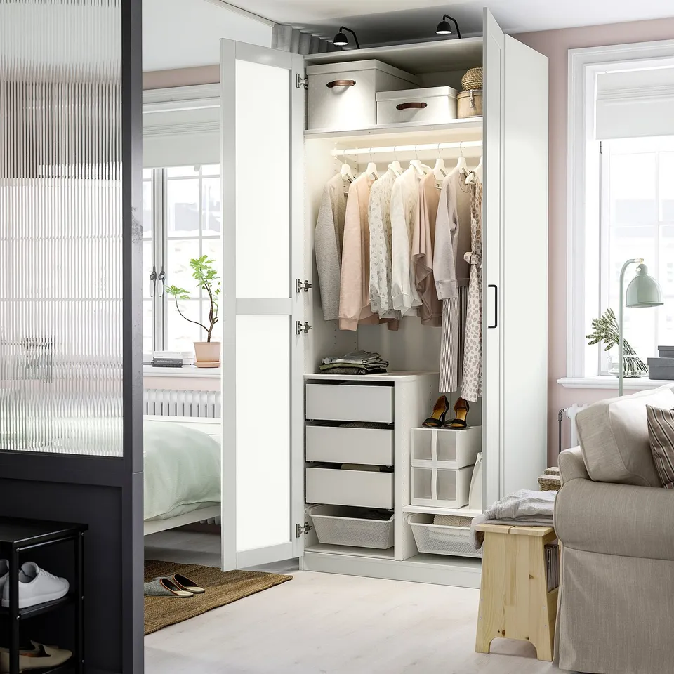 Open wardrobe with clothes, drawers, and baskets inside, next to a bed.