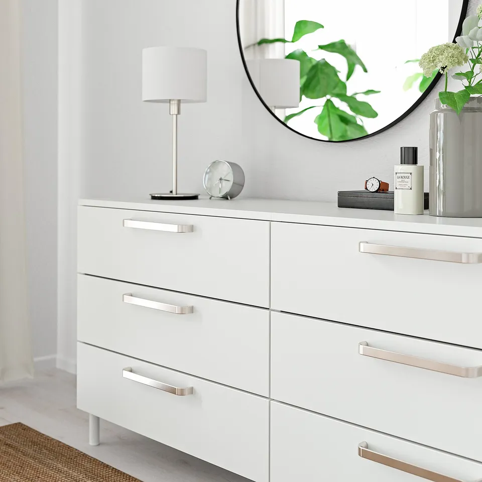 White dresser with 6 silver-handled drawers, topped with lamp, clock, mirror, and decor.