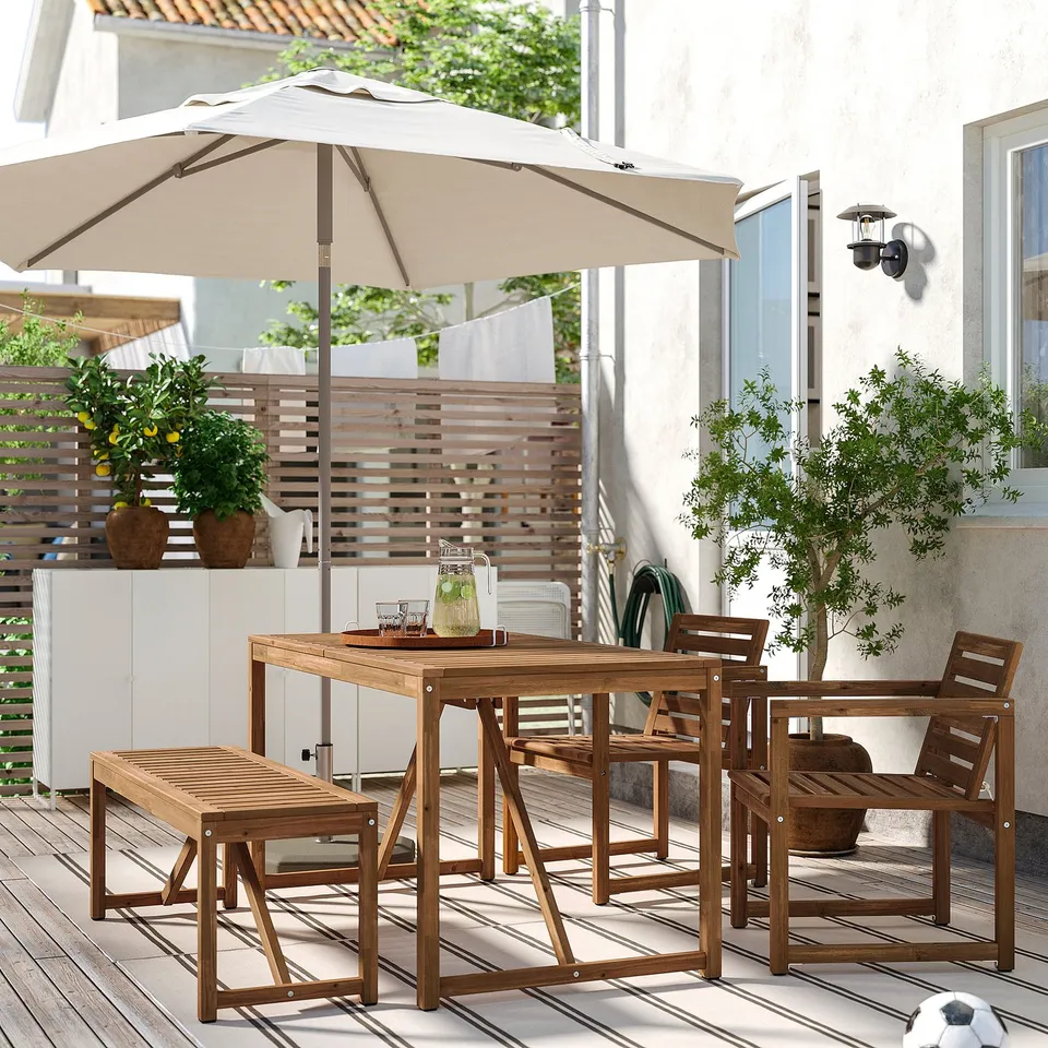 Wooden patio set with table, two chairs, bench, and umbrella. Potted plants, white building in background.