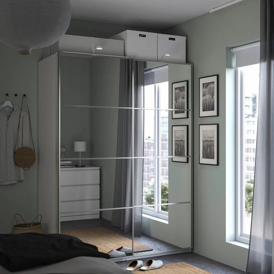 Modern bedroom with PAX wardrobe, mirrored doors, mixed storage. Sheer curtains, art, shirt on hook, small bedside lamp.