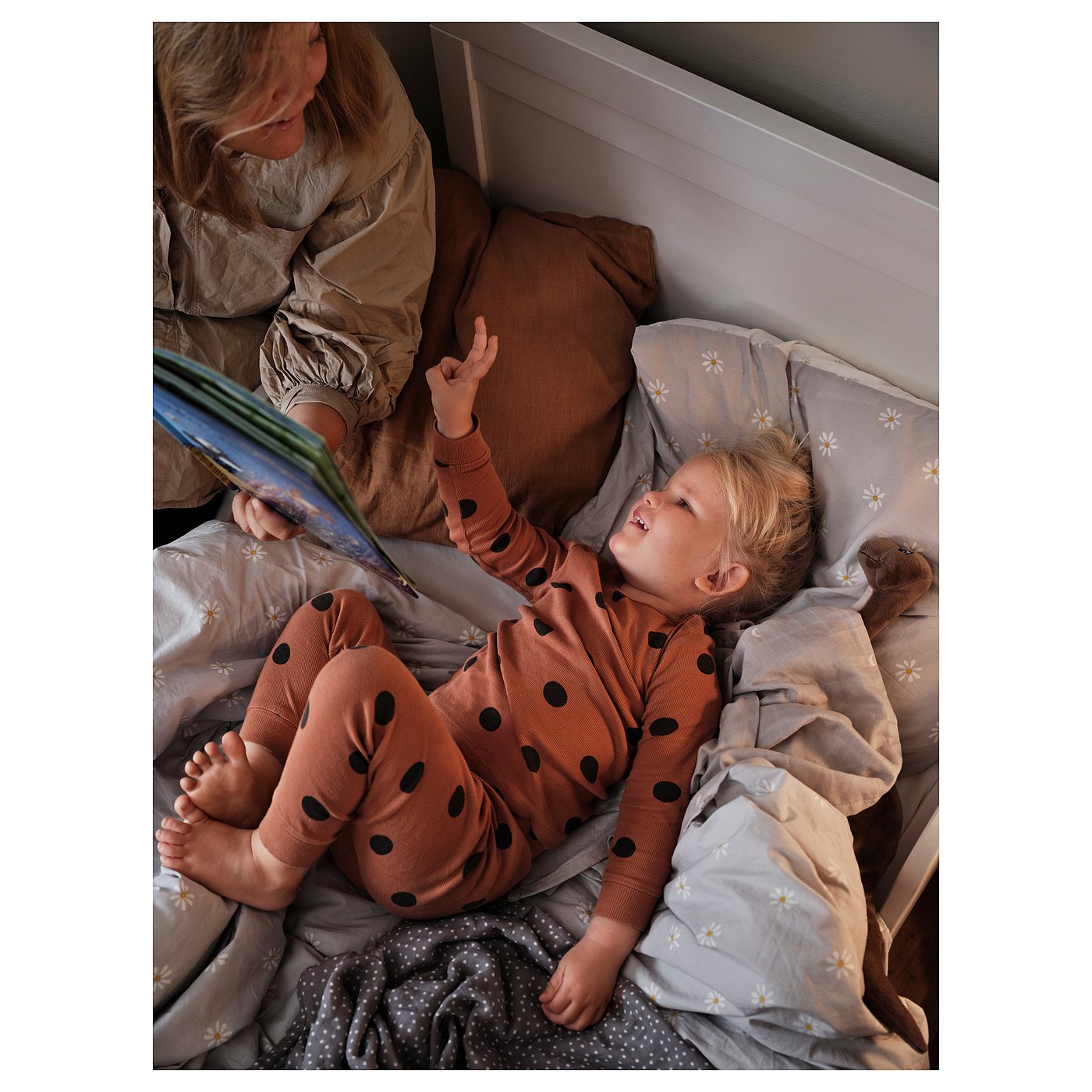 Two children read a book in bed, under a floral-patterned grey duvet.