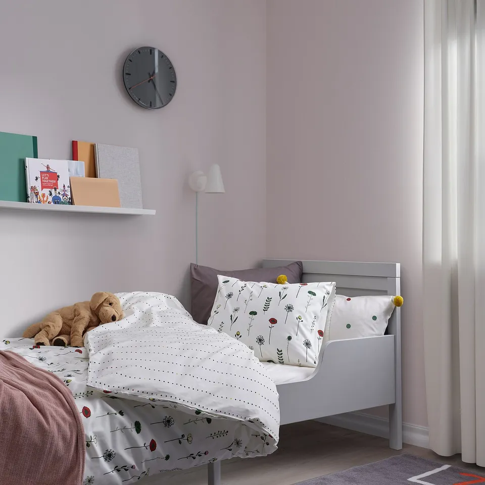 Kids bed with NATTSLÄNDA lavender duvet, floral patterns. Against lilac walls, with wall clock, bookshelf, lamp, and teddy bear.