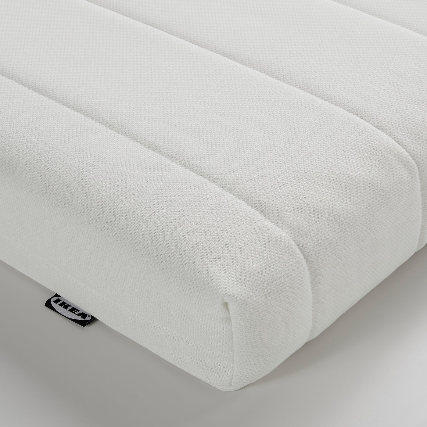 Close-up of white foam mattress, smooth texture, neatly folded corner. Brand label visible.