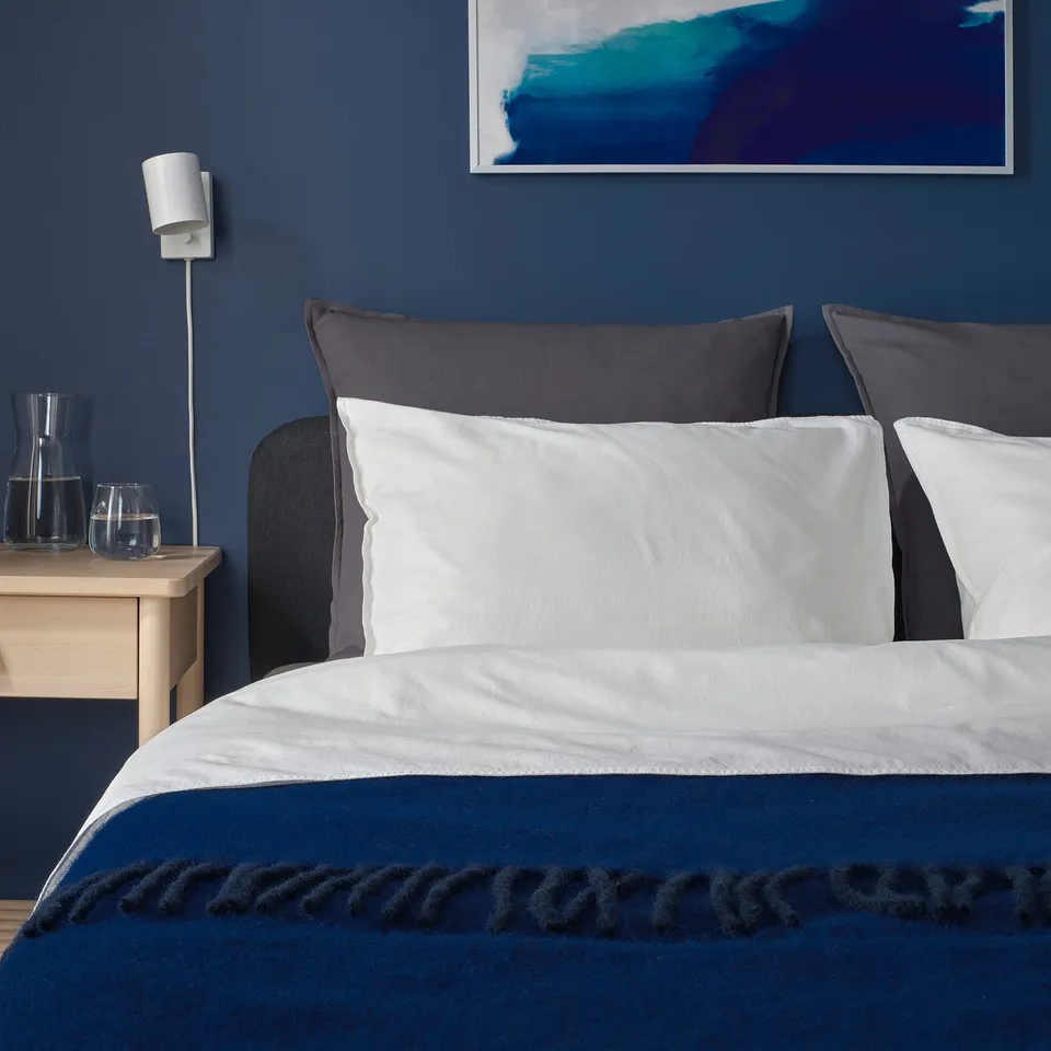 Bed with large grey pillow & blue throw; minimalist bedside table, lamp, and art.