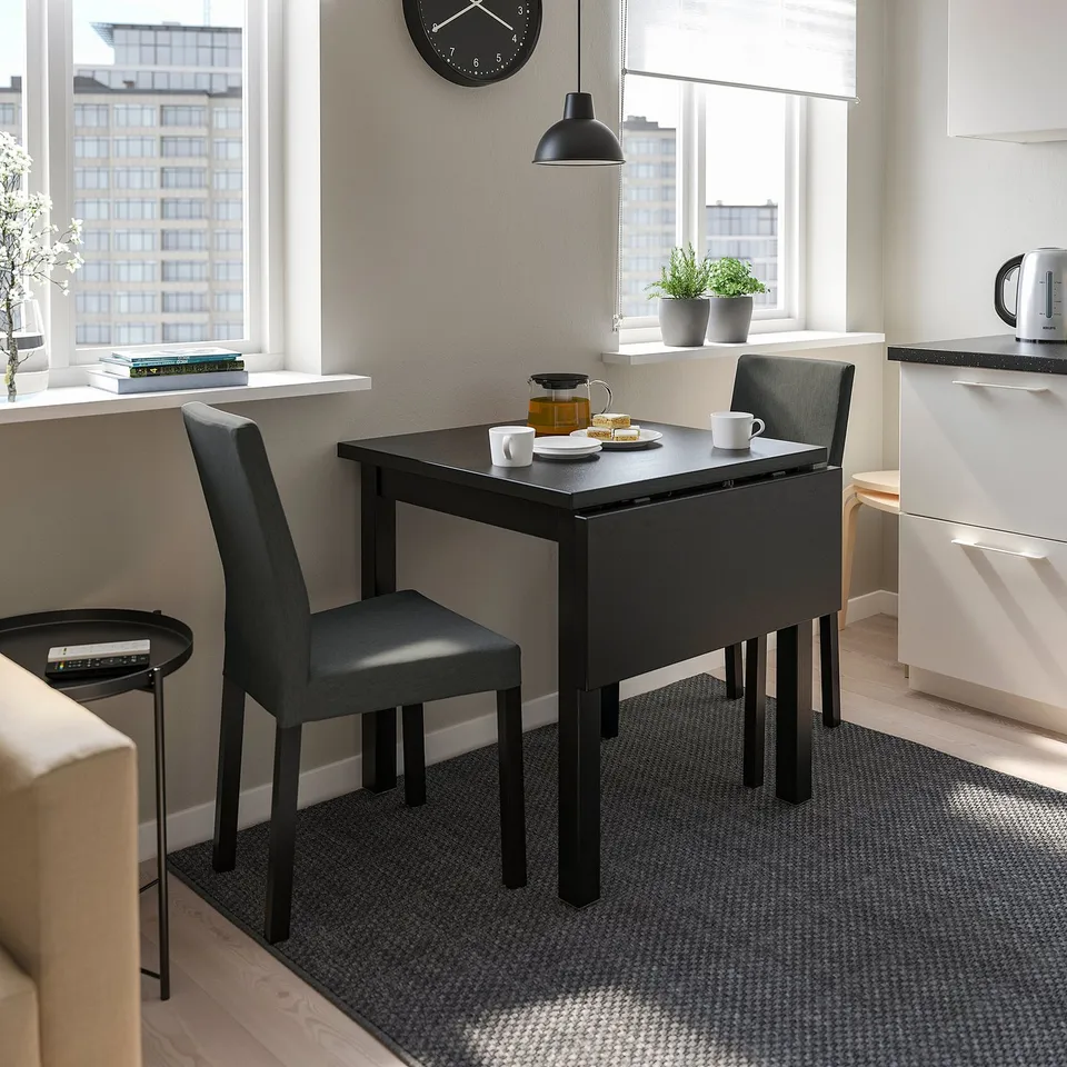 Modern black drop-leaf dining table with two padded chairs. Compact design ideal for small spaces. Large windows offer city views.