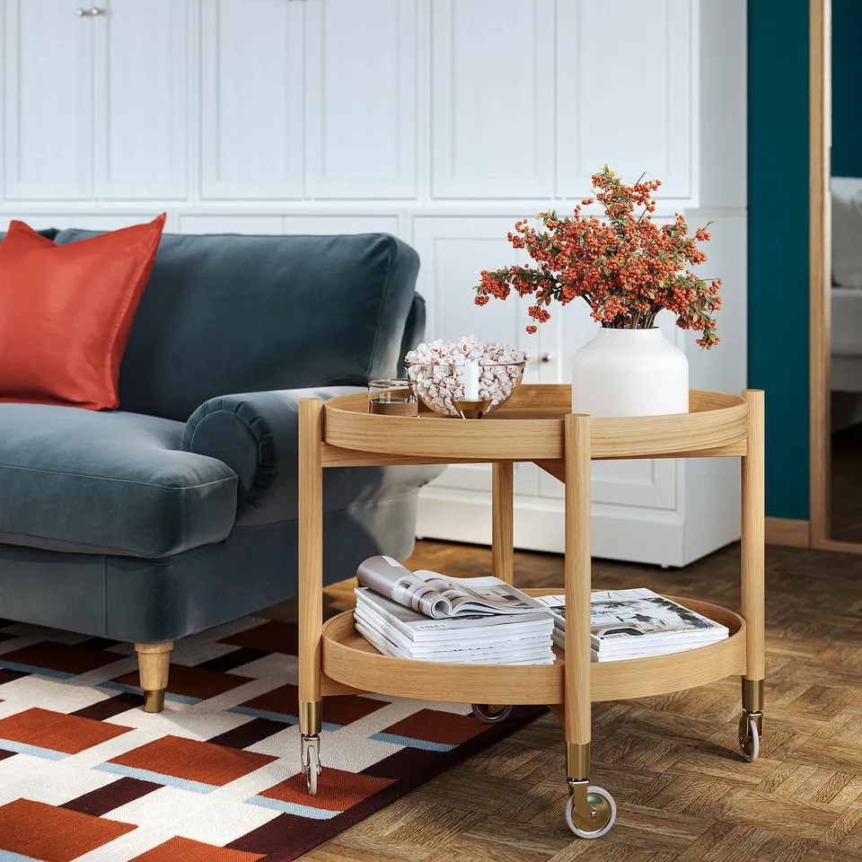 Valnas light oak serving trolley with brass details and removable tray. Next to blue couch, showcasing orange flowers in a white vase on top.