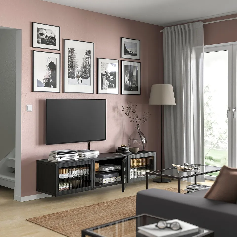 Modern living room with BESTÅ tv stand, pink walls, grey curtains, photos, wooden floor, glass coffee table with books.